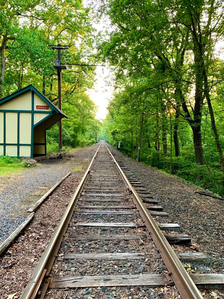 A view of train tracks extending straight into the woods under a partly cloudy sky. A small wooden train station, painted light yellow-beige, stands on the left.