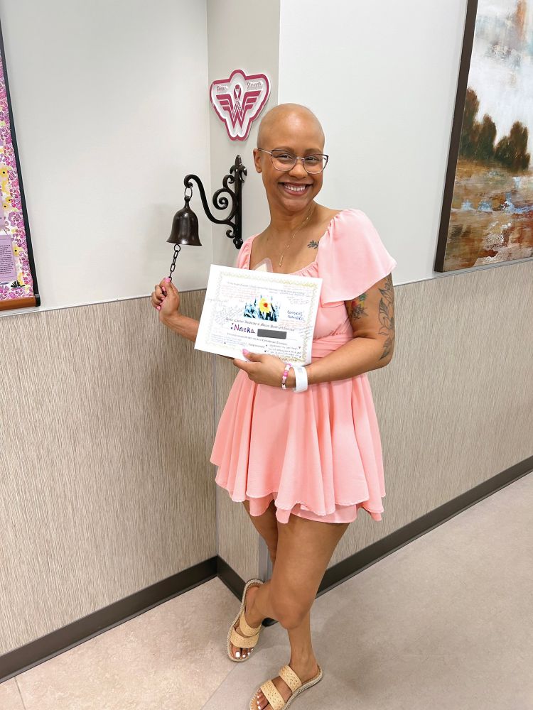 neeks, wearing a pink dress and holding a completion certificate, smiling and ringing the chemo bell