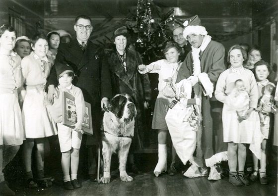 black and white photo of a group of children and hospital managers standing in front of a Christmas tree,, Santa is handing out presents to the children. Bonus St Bernard dog watches on