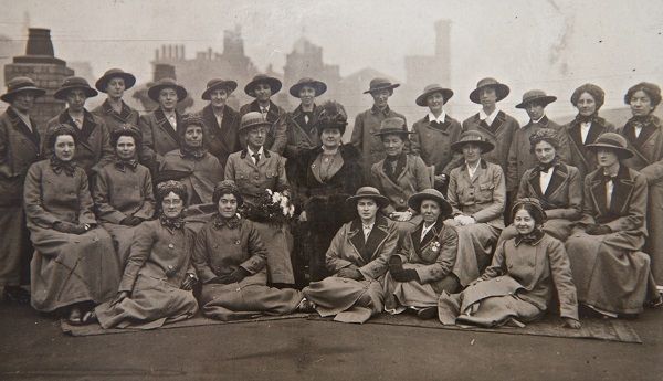 Black and white photo of a group of women. Arranged in three rows the front row sits on the ground, the second on chairs and the third standing. They are all wearing long overcoats and hats. Chimneys can be seen in the background.