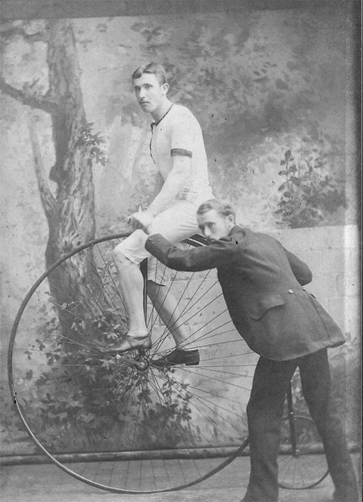 Black and white photograph of two men and a penny farthing bicycle. One man is sitting on the penny farthing and the other man is standing next to it, supporting the bike.