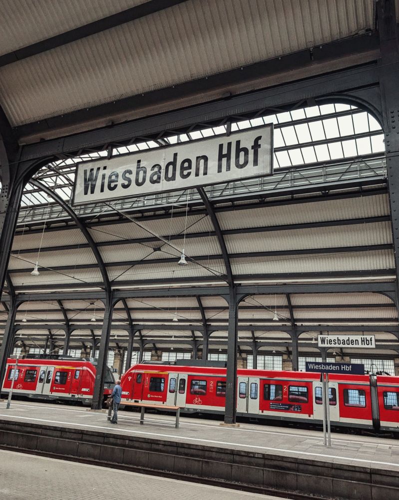 A photo of the old Wiesbaden Hbf (Hauptbahnhof) signs suspended above the train platforms. There is a red and white train visible three platforms away. 