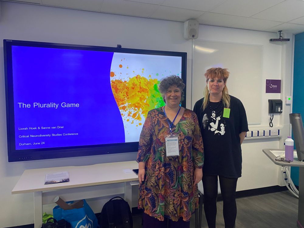 Liorah Hoek and Sanne van Driel smiling at the camera. They are standing in front of a PowerPoint slide in a seminar room. On half of the slide is blue and the other is white with orange and green paint splotches. On the blue slide of the slide the text reads 'The Plurality Game'. 