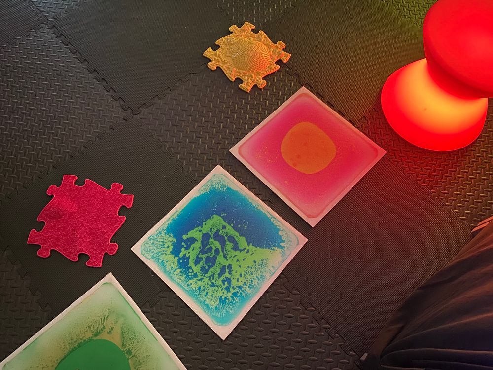 The floor in The Sensory Den – a pop up sensory room. There are yellow and red jigsaw pieces, a red glowing stool, and three marble effect tiles in pink and orange and green and blue. 