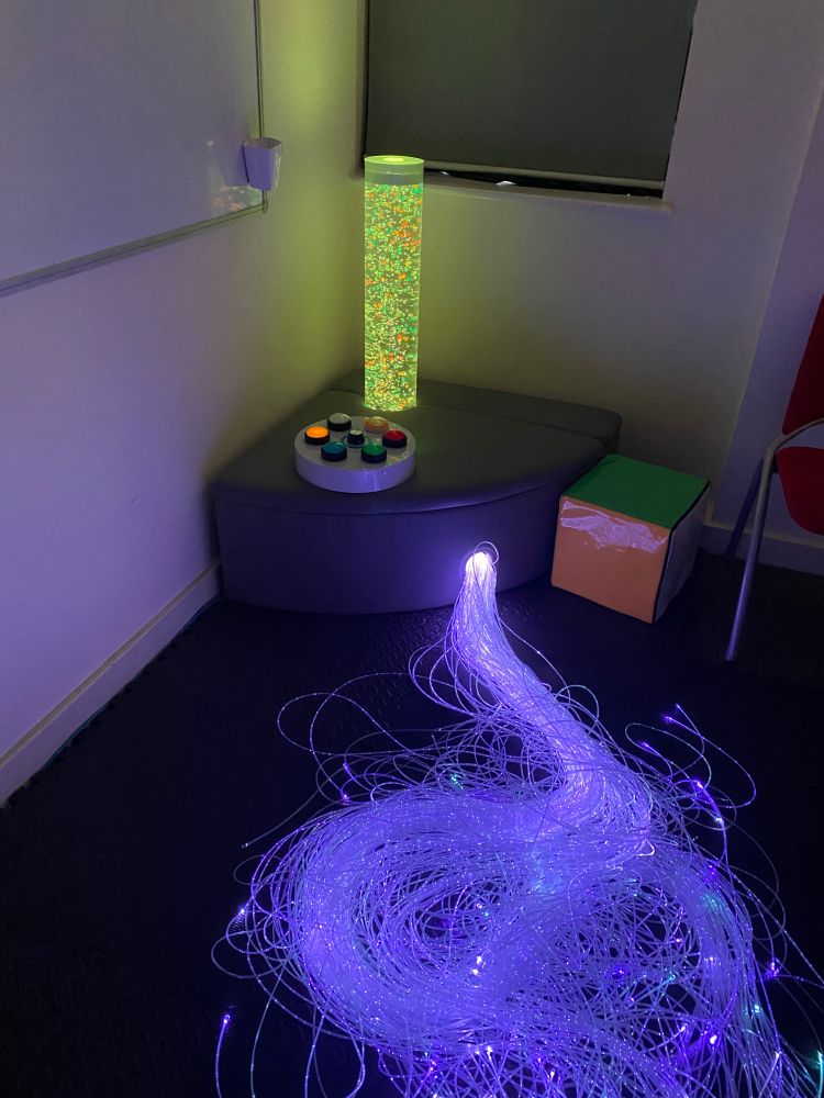 A lime green and orange lava lamp on a bench, a large cube to the right, and fibre optic tails in white/light blue on the floor.
