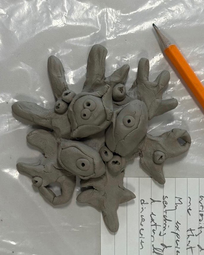 A clay sculpture with round eye-like shapes on a plastic sheet. There is an orange lead pencil and a card with hand-writing on it to the right hand side. 