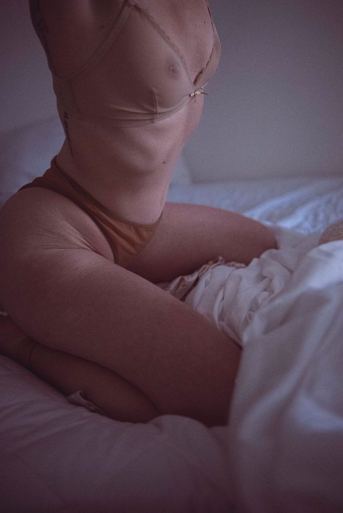 A high grain color photo of a feminine body kneeling, posed, on a bed. Their pelvis and spine and shoulders are organized and stacked, creating length in their waist, a narrowness over rounded hips. Their knees are spread, their underwear riding their lines. 