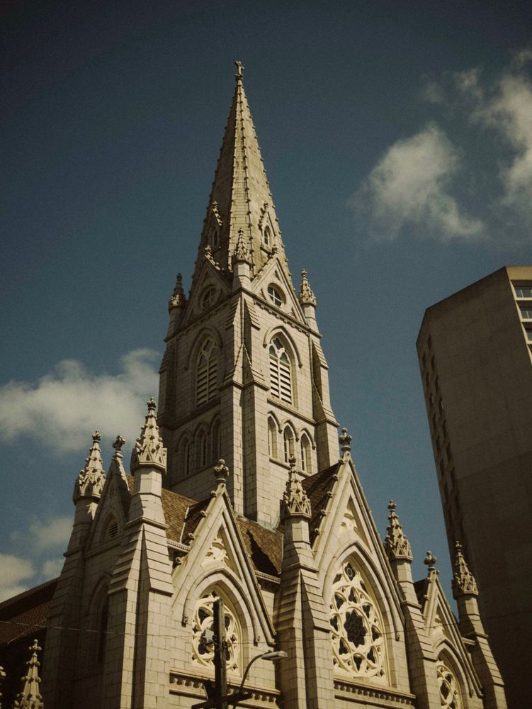 in the hot blue sky are two grey structures, one is the ordinate basilica spire filling the center of the image, gothic and oppressively beautiful it almsot overflows the image, crowding out the glimpse of a brutalist skyscraper to the right. 