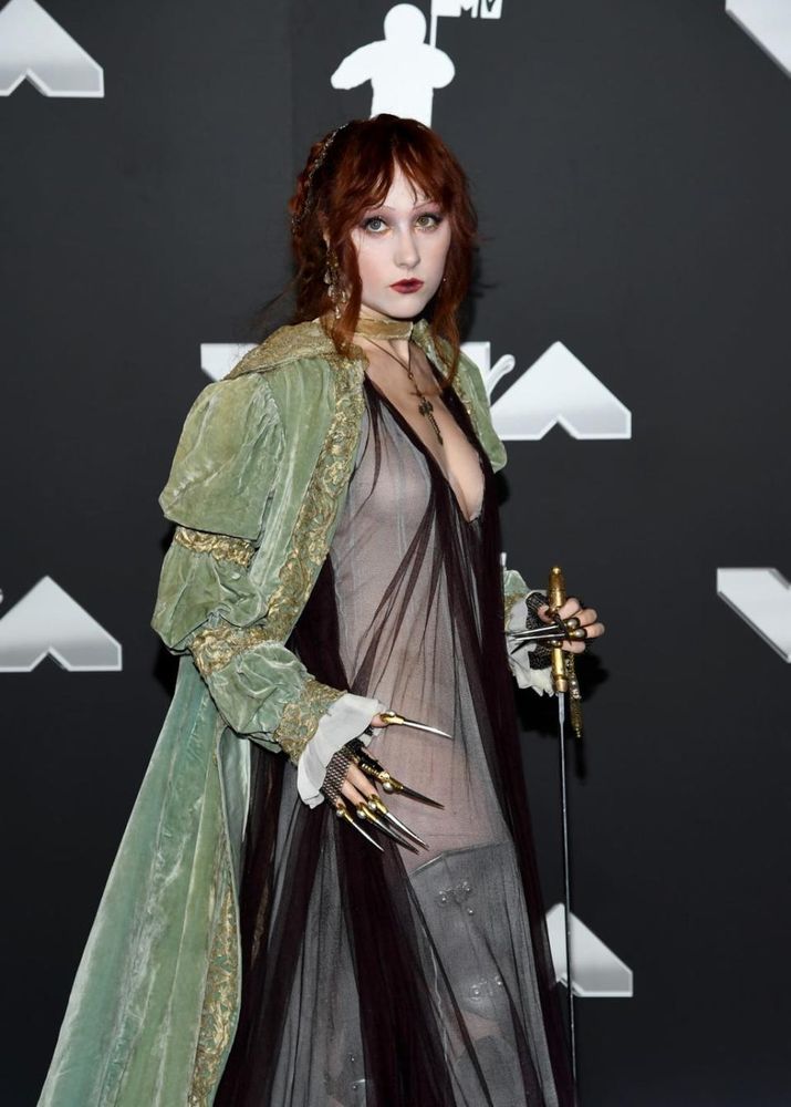 Chappell Roan attended the 2024 MTV awards in a pistachio velvet robe with gold embroidery along the lining, collar, and puffy, ruched sleeves.

Underneath, she wore a sheer gown with mid-length sleeves and a plunging neckline. Her accessories included leather bracers featuring chainmail wrist cuffs, armored rings with inches-long metallic nails, steel plate legs, a cross amulet, and a longsword.