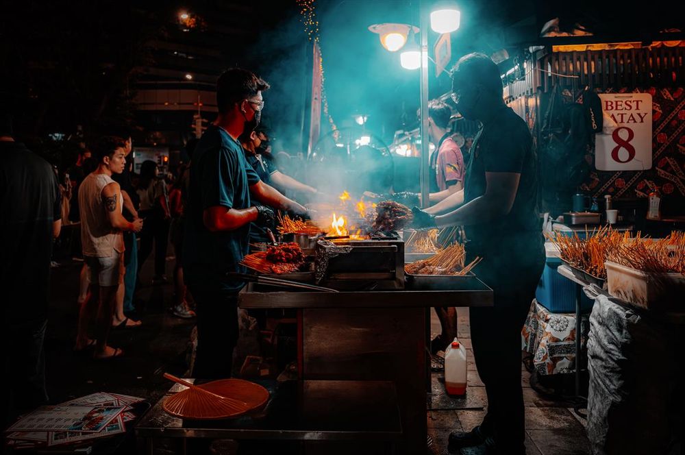 A dark scene filled with smoke focused on two streetside vendors barbequing chicken and mutton skewers. 