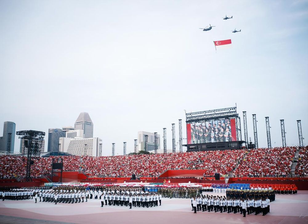 Singapore's national day parade at the Padang. Contingents are formed up with three helicopters flying a large national flag in the background.