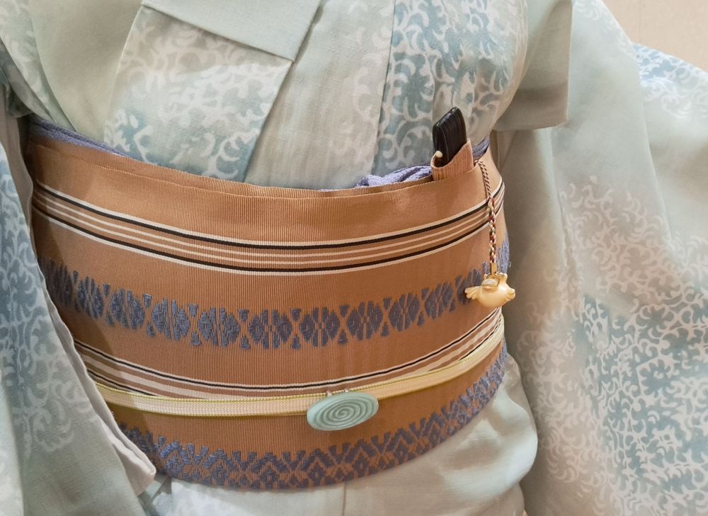 水色濃淡に白抜きの唐草をあしらった地に千鳥が飛ぶ柄の着物だが写真に千鳥は写っていない。茶に青と白の博多献上帯