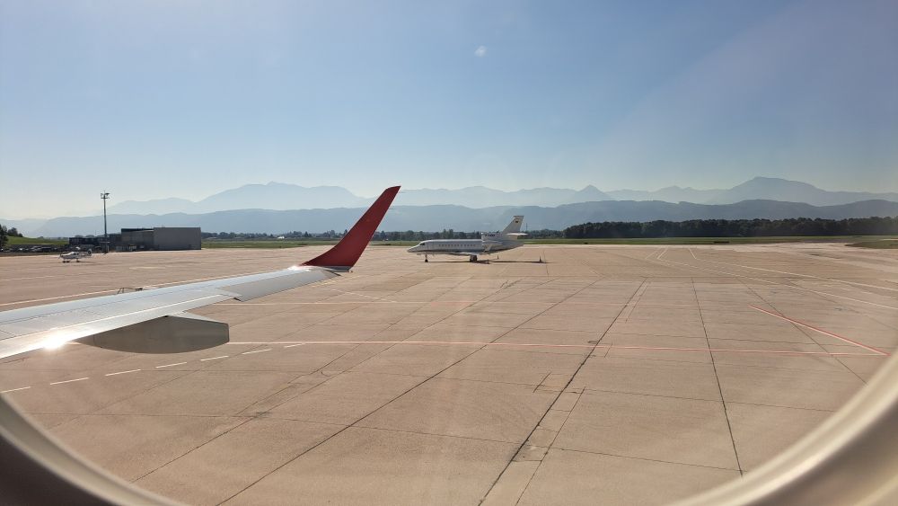 Shot through airplane window showing a bit of a wing, a tiny plane, and mountains in the distance