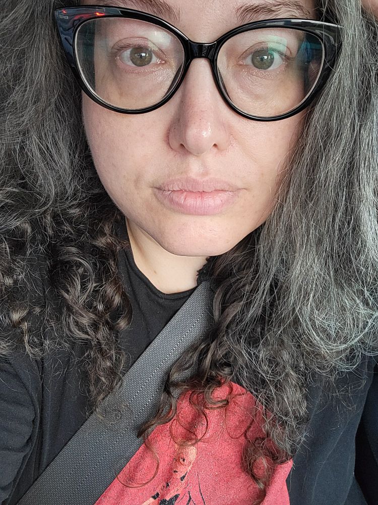 A white woman with deep eyelashes and black glasses. She has wavy Grey and brown hair and a bland expression 