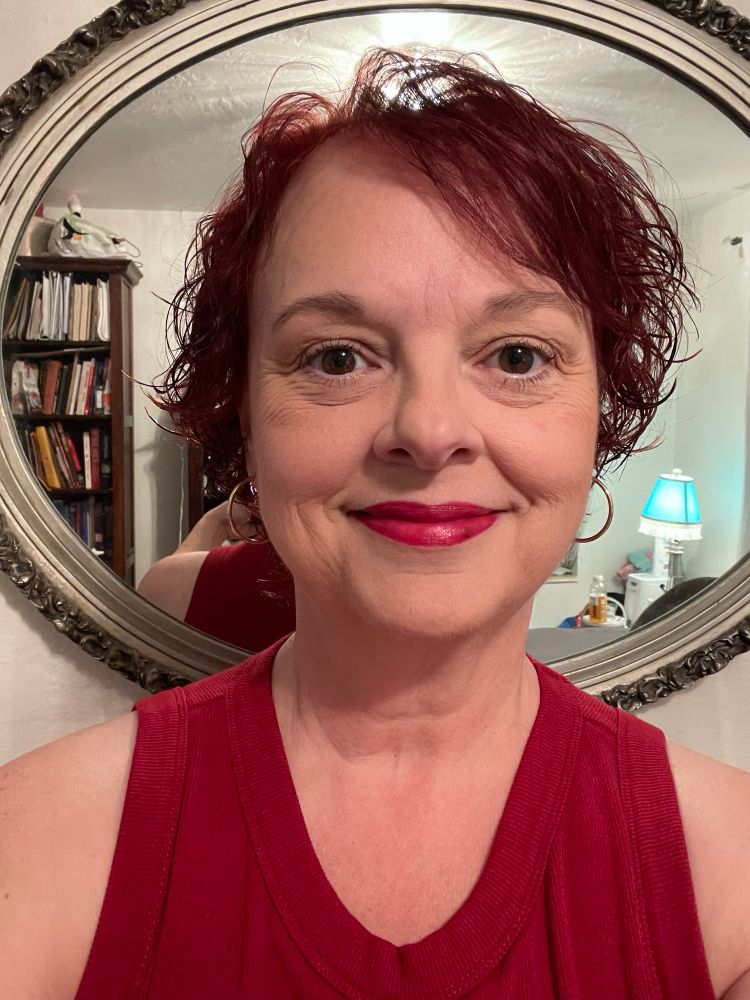 Original poster posing for selfie with newly coiffed hairdo, short and curly and deep auburn in color, makeup applied with a reddish lip. Maroon sleeveless top. Standing in front of a mirror. 