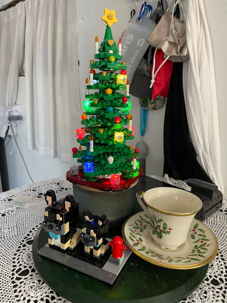 A table centerpiece display of a LEGO Christmas tree and a LEGO large and small French Bulldog. The display is on a round table with a crocheted tablecloth. There is a Lenox Holiday pattern cup and saucer next to the LEGO sets. The latter is green holly leaves and red berries on a cream background. The cup and saucer have gold trim around the rims. 