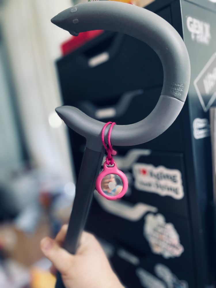 A hand is holding a gray cane with a curved handle. Attached to the handle is a pink keychain with a circular reflective surface. In the background, there is a black drawer unit with various stickers, including one that reads 'I Love Aging and Dying.’
