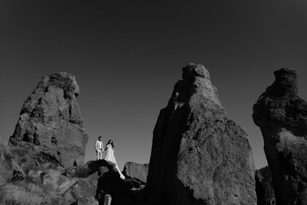 Black and white image of bride and groom standing between tower rocks.