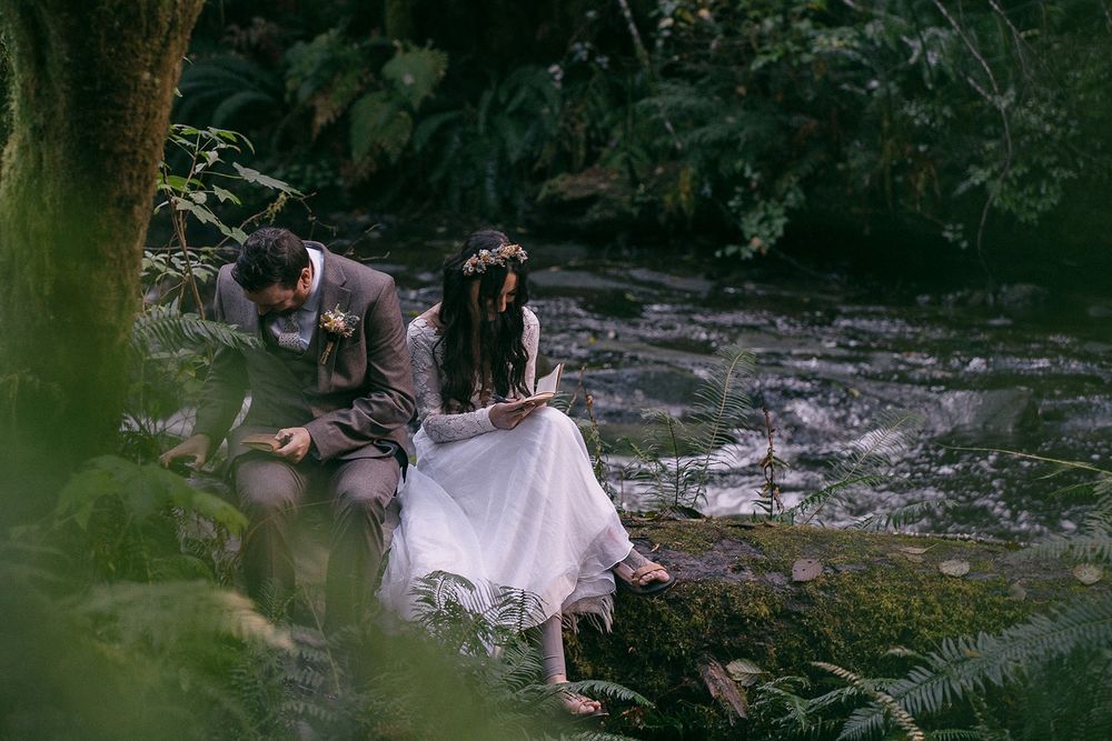 A bride and groom sitting on a log by a stream, with their heads bent low, writing in small journals in their laps. 