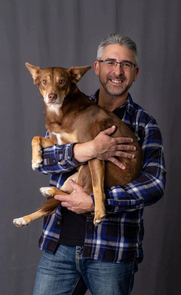 Me, standing and smiling in jeans and a purple-and-black plaid shirt, holding my handsome brown dog Carson, whose facial expression is that of both quiet stoicism and a hint of "I love you, but please put me back down".