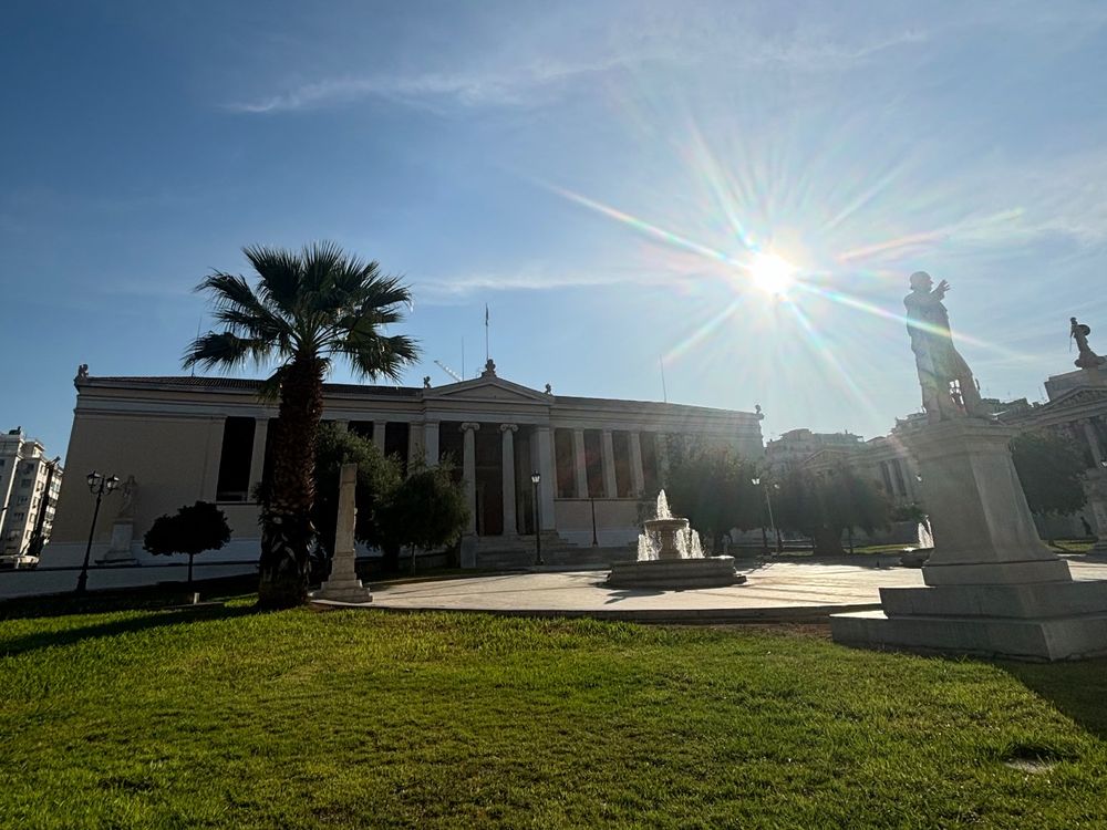Landscape of University of Athens facade. There is a garden in the front with a fountain, a statue and some trees. The sky is clear and the sun is bright low at the upper right corner