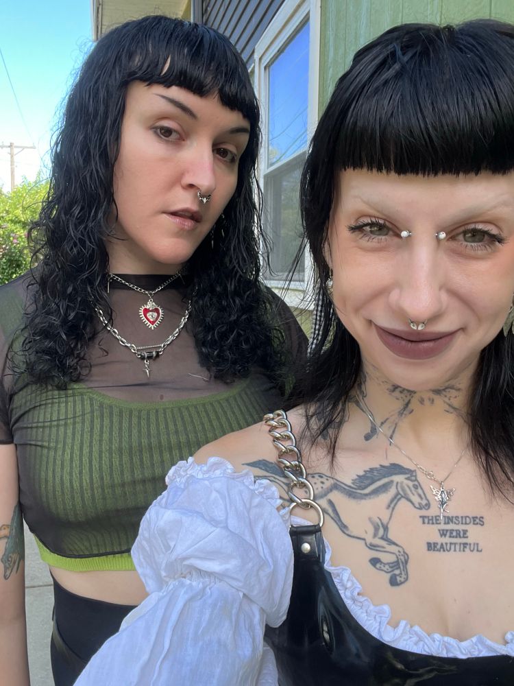 Pony, a thin white femme with tattoos and facial piercings wearing a white top with latex over it, Dolly stands behind her wearing a black sheer top with a green tank underneath, she is taller and her hair is curly.