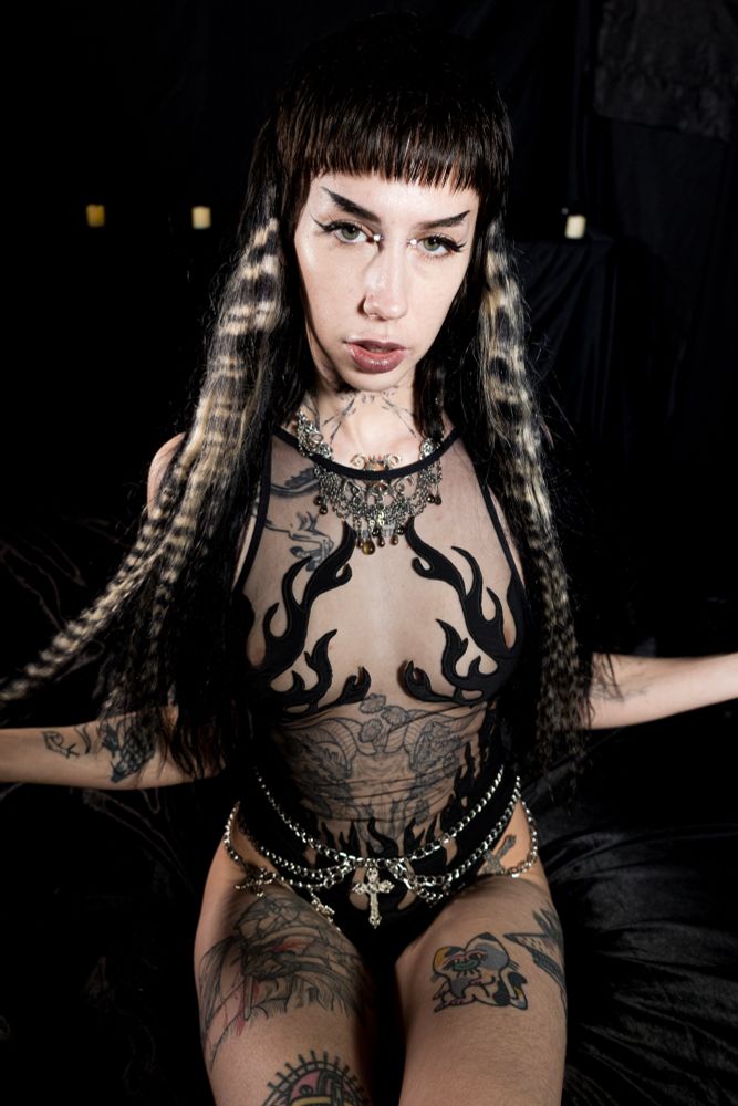 Pony sits on a black silk bed wearing a black flame print bodysuit, chain belt and chainmail necklace with raccoon tail extensions 