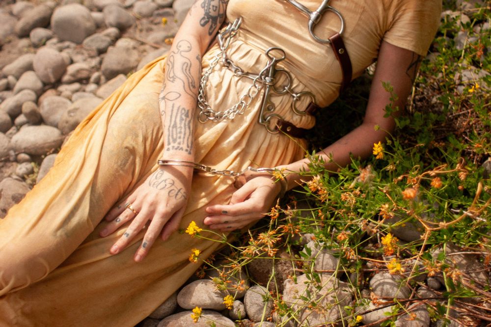 Pony wears a cream colored dress soaked in river water with horse-bit harnesses strapped to her chest, hands cuffed in metal cuffs, yellow flowers sparkle beside her