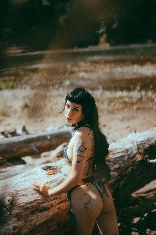 Pony leans on a fallen log by the lake, long ponytail and metallic bikini, she looks over her shoulder at the camera 