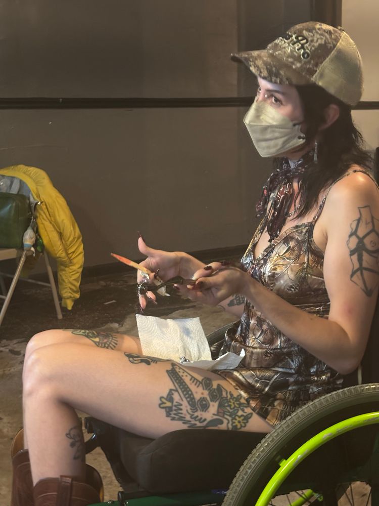 Pony wears a face mask, baseball cap and Realtree slip dress with cowboy boots, they are in a green wheelchair, they’re holding 2 knives 