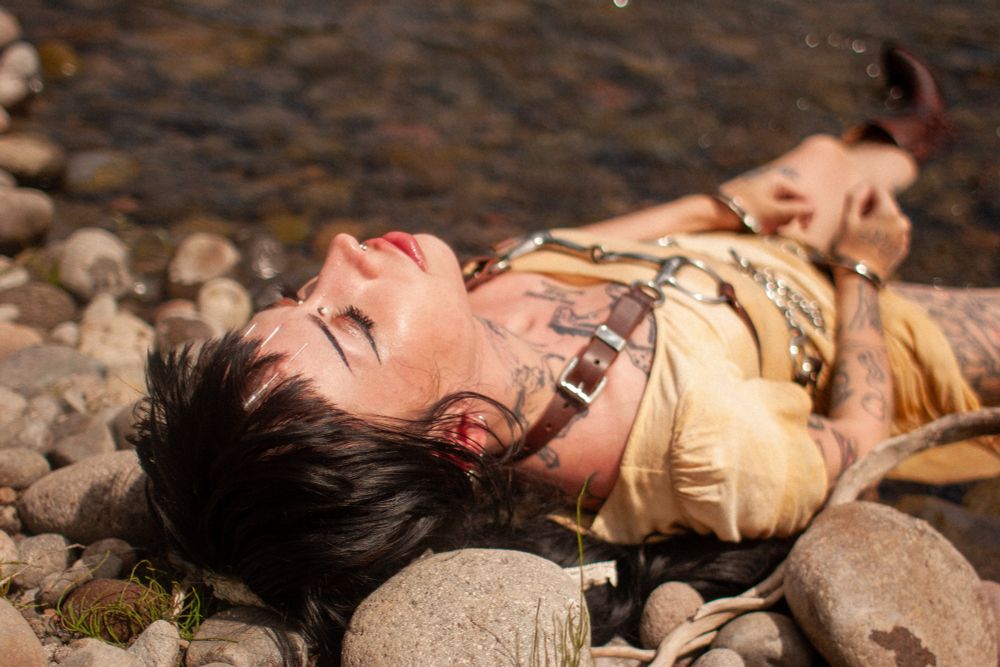 Pony lays her head back on a rocky river shore, needles protruding from her forehead as the sun hits her face 
