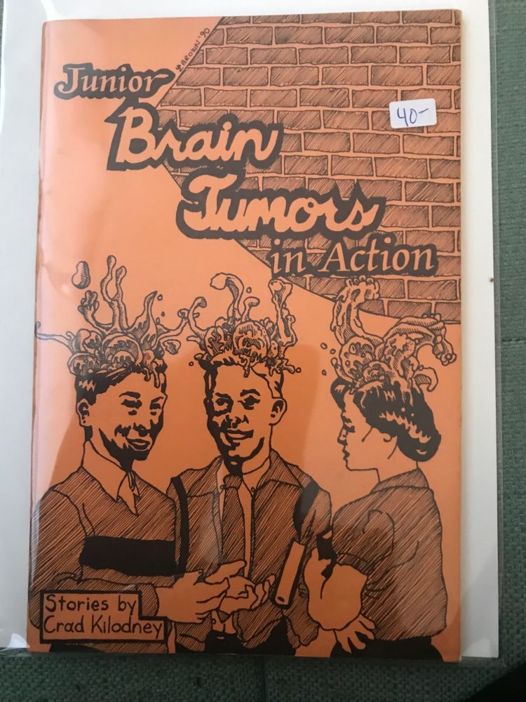 Junior Brain Tumours In Action by Crad Kilodney, 1990. The cover is orange cardstock with black printing. Two youg men and a young woman stand clustered together, smiling, giant amoeba-like tumours erupting from the tops of their heads. 