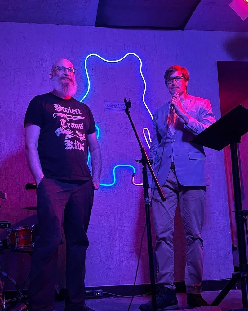 Co-editors of Devouring TOMORROW, A.G. Pasquella and Jeff Dupuis, on stage at Teddy Beer for the Toronto launch of DEVOURING TOMORROW. Jeff is looking dapper in a blazer and A.G. is wearing a black T-Shirt that says, “Protect Trans Kids.” 