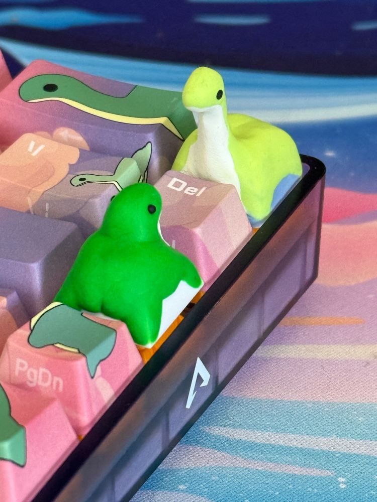two nessies keycaps looking at each other on a nessie themed keyboard (from apex legends) - one has a shorter neck and is looking up at the other one