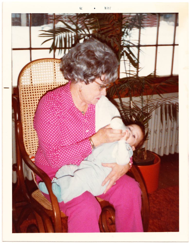 A woman (maternal grand mother) in a very 70s pink outfit holding a baby (moi) and bottle feeding it.