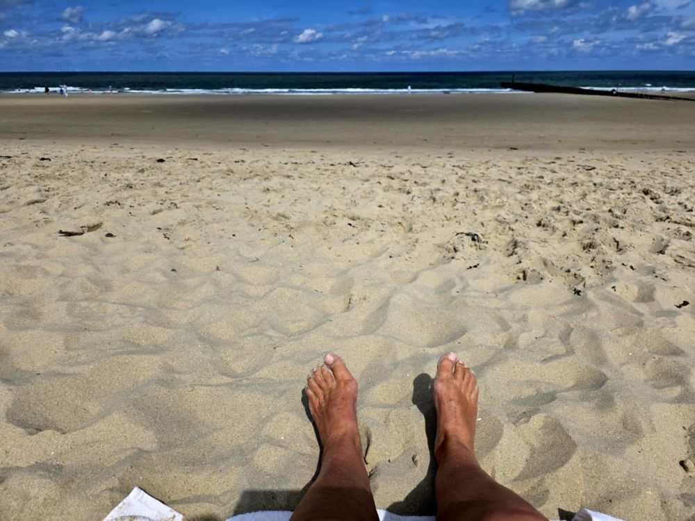 Im unteren Bildbereich sind die braungebrannten Unterschenkel und Füße einer Person zu sehen, die im feinen gelben Sand am Strand sitzt, in einigen Metern Entfernung schlägt das dunkelblaue Meer mit sanften Wellen an den Strand, am blauen Himmel ziehen ein paar Wolken.