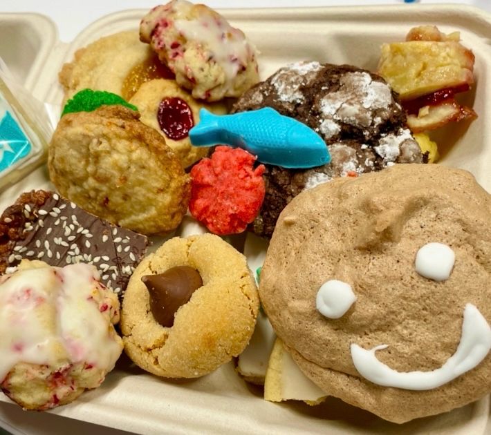 Paperboard takeaway container brimming with assorted cookies. Front and center is a big round one that’s pale brown with a frosting smily face on it. On top is a sky blue molded candy fish.