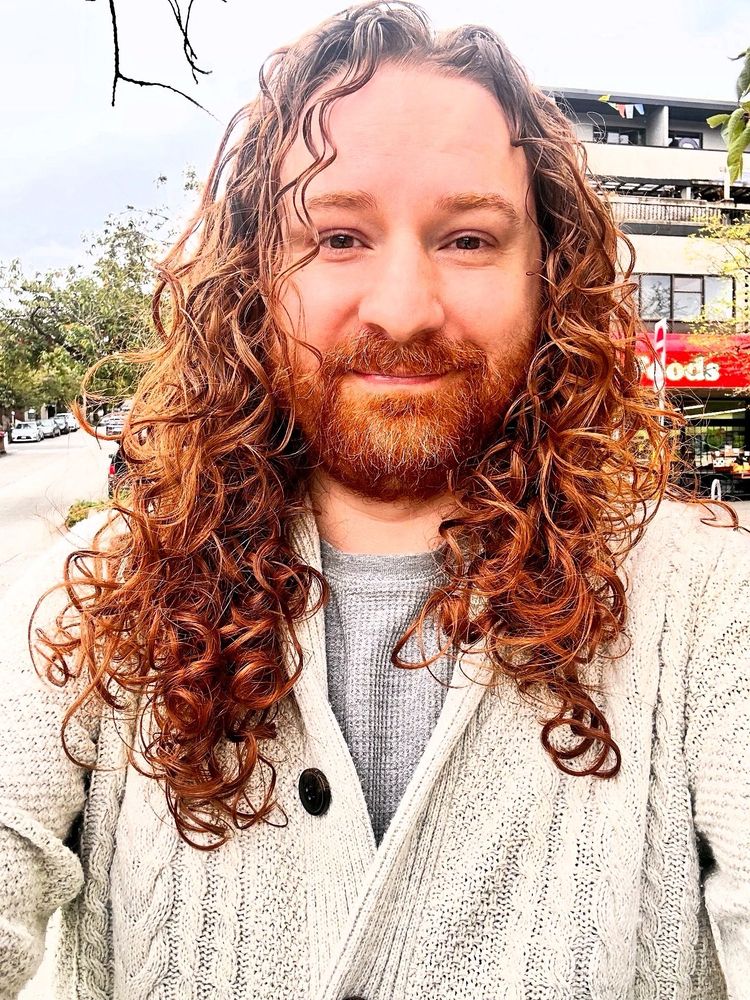 Morgan wearing a white cardigan, standing outside, notably with very curly shorter but still past the shoulder red hair 