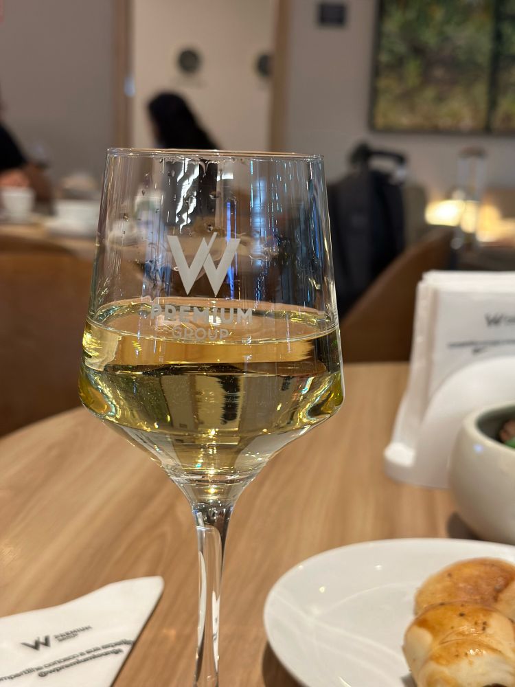 Uma taça de vinho branco é capturada em primeiro plano, em modo retrato, com detalhes nítidos e cores vibrantes. O foco da imagem está no W gravado no vidro da taça, uma referência ao W Premium Group, que administra a sala VIP onde a foto foi tirada. O vinho brilha sob a luz suave, destacando a sofisticação e a exclusividade do ambiente.