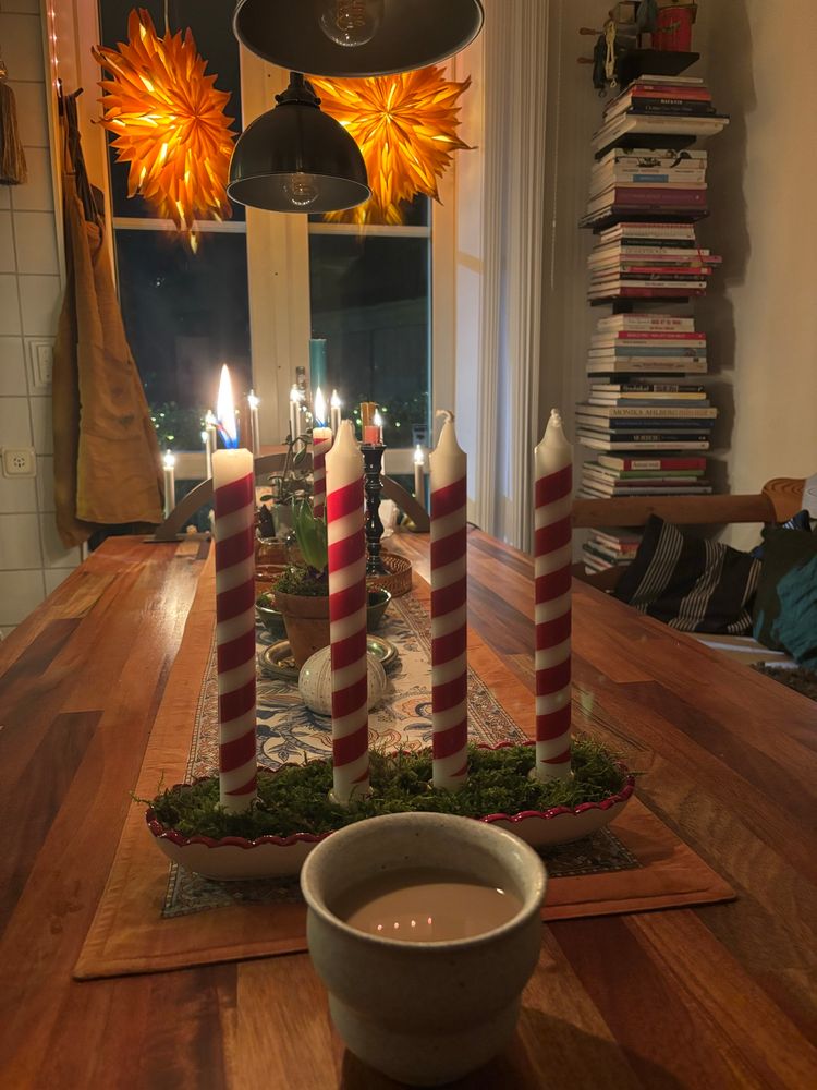 Ett köksbord med en kaffekopp närmast kameran, och bakom den en adventsljusstake med mossa och 4 rödvita ljus varav det första är tänt. Längst bort på bilden ett fönster med 2 orange julstjärnor varav den ena såklart hänger snett. 🙄
