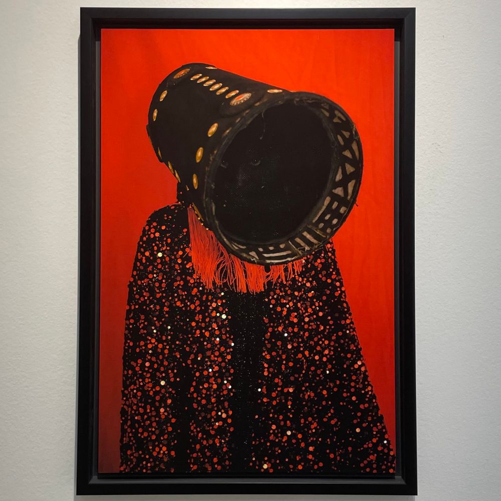 A framed photographic print of a figure wearing a robe of hand-stitched red and black sequins, a black rigid conical shroud hides the face within, but a close inspection reveals a feline face inside. 