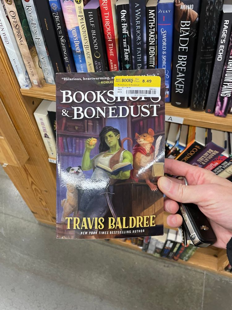 A paperback of Bookshops & Bonedust, by Travis Baldree, held in a right hand, along with some car keys. Behind are a few bookstore shelves.