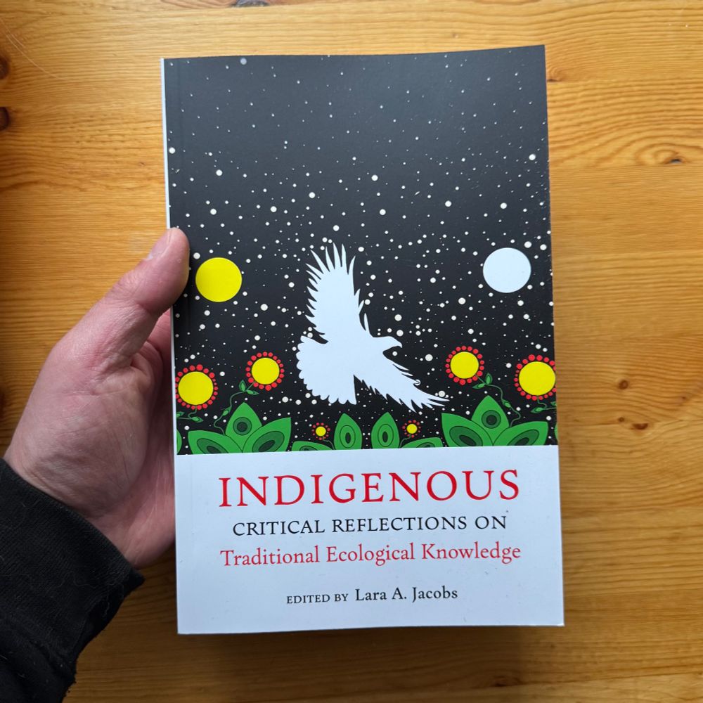 a hand holding up the paperback book, "Indigenous Critical Reflections on Traditional Ecological Knowledge". on the cover is an eagle silhouette in white in front of a starry background and above green stylized flower leaves and yellow flowers with red petals.