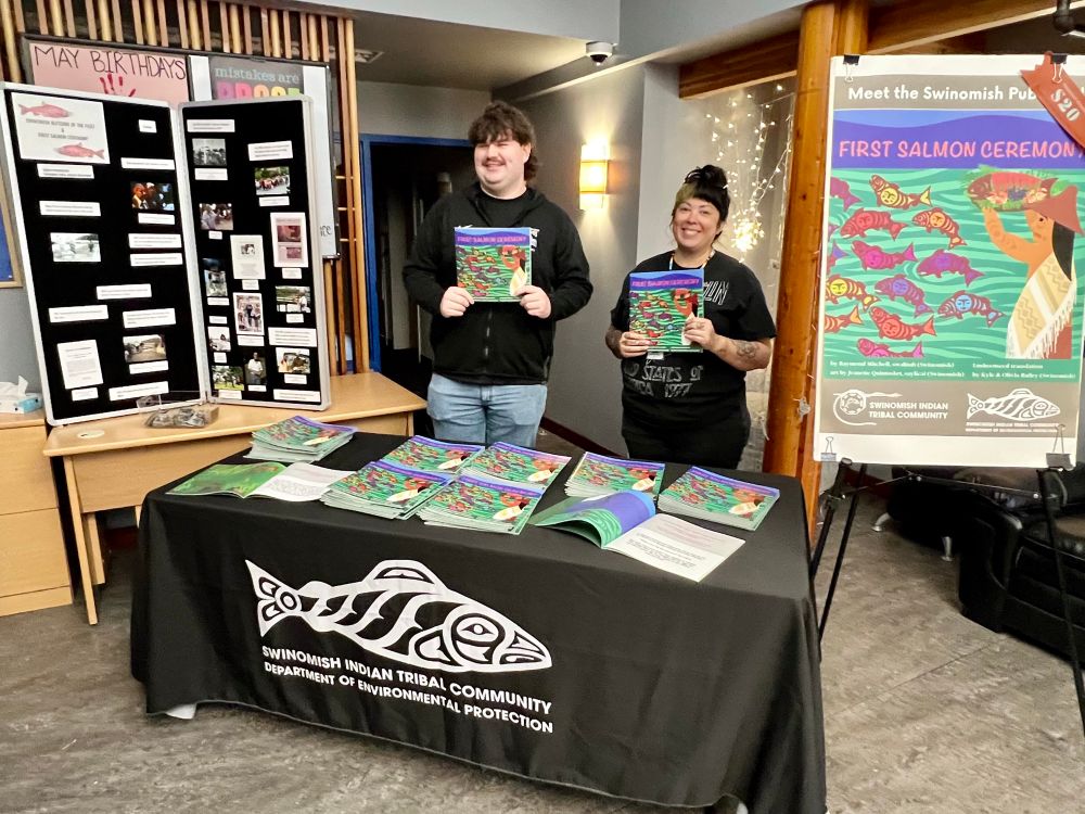 Two people holding up 1st salmon ceremony books standing behind a table with stacks of the books. to one side is a display board with photos and captions of past 1st salmon - blessing of fleet ceremonies, to the other side is a big poster of the 1st salmon ceremony books standing behind cover.