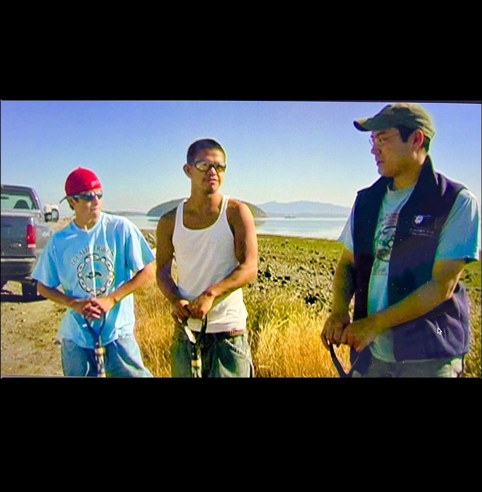 Swinomish Tribal youth filmmakers Cody ( behind camera), Travis, Nick and Todd on a beach, holding clam forks on a sunny day in summer.