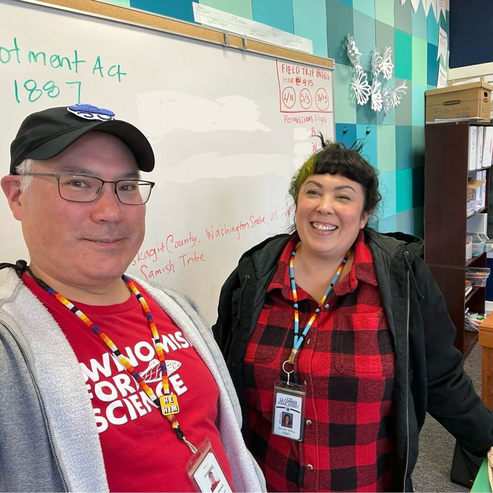 Selfie with Swinomish Indigenous science teachers Todd and Jen Willup wearing beaded lanyards with school IDs and standing in front of a whiteboard.