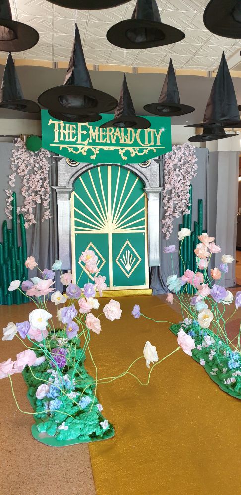 A display for the Wicked movie. Black witches hats are suspended from the ceiling over a gold carpet leading towards a green, gold, and silver door. A sign above the door reads "The Emerald City". Artificial flowers hang next to the entryway and are also placed along the gold carpet.