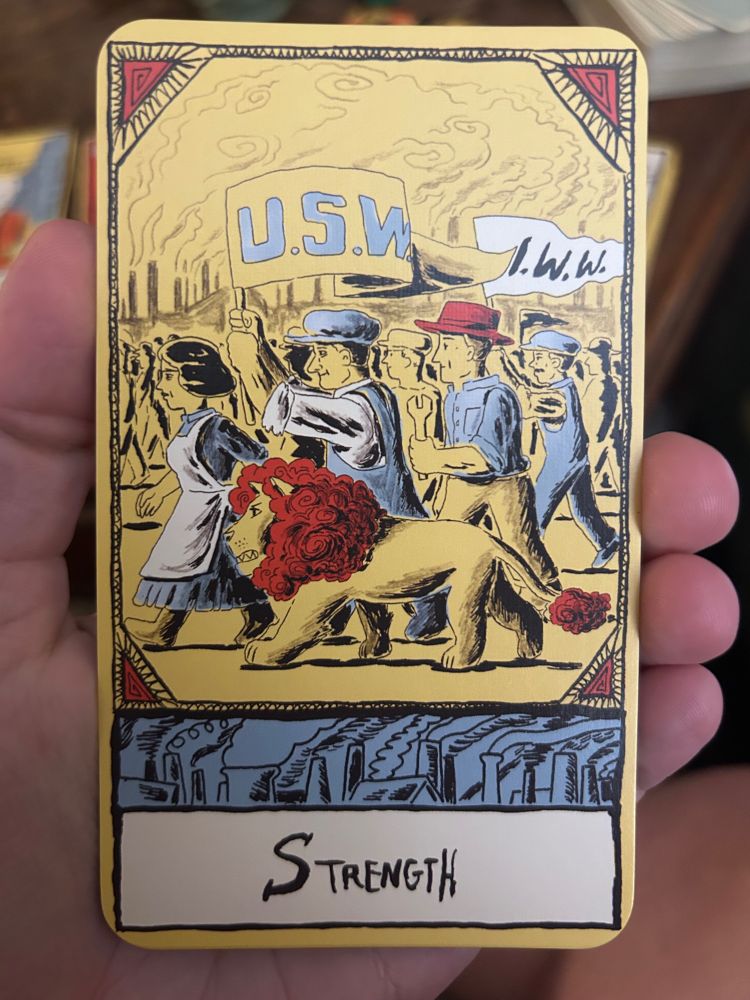A tarot card titled “Strength” from The Pittsburgh Tarot, depicting a woman walking beside a lion with a red mane amid a crowd of industrial workers carrying union banners labeled “U.S.W.” and “I.W.W.” Smoke rises from factory chimneys in the background.