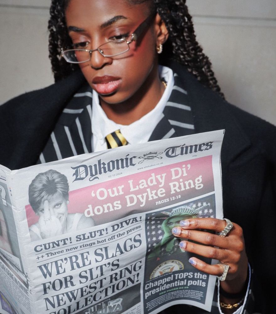 cat burns in a suit holding a newspaper from the “dykonic times” with the headline “‘our lady di’ dons dyke ring”