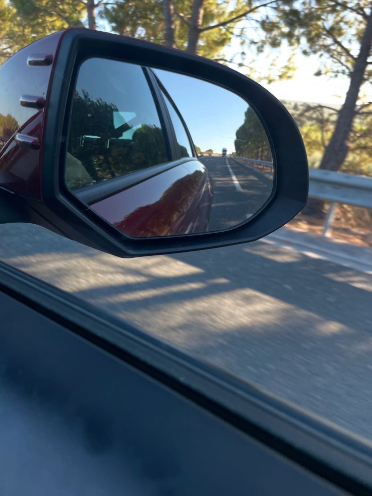 Foto desde la ventana del coche, se ve el retrovisor y parte de la carrocería roja del coche. Carretera y arbolitos.